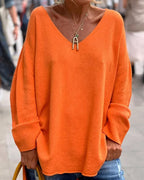 Femme portant un pull oversize orange vif avec un col en V, accessoirisé d'un collier doré en forme de lettre, tenue décontractée en extérieur.