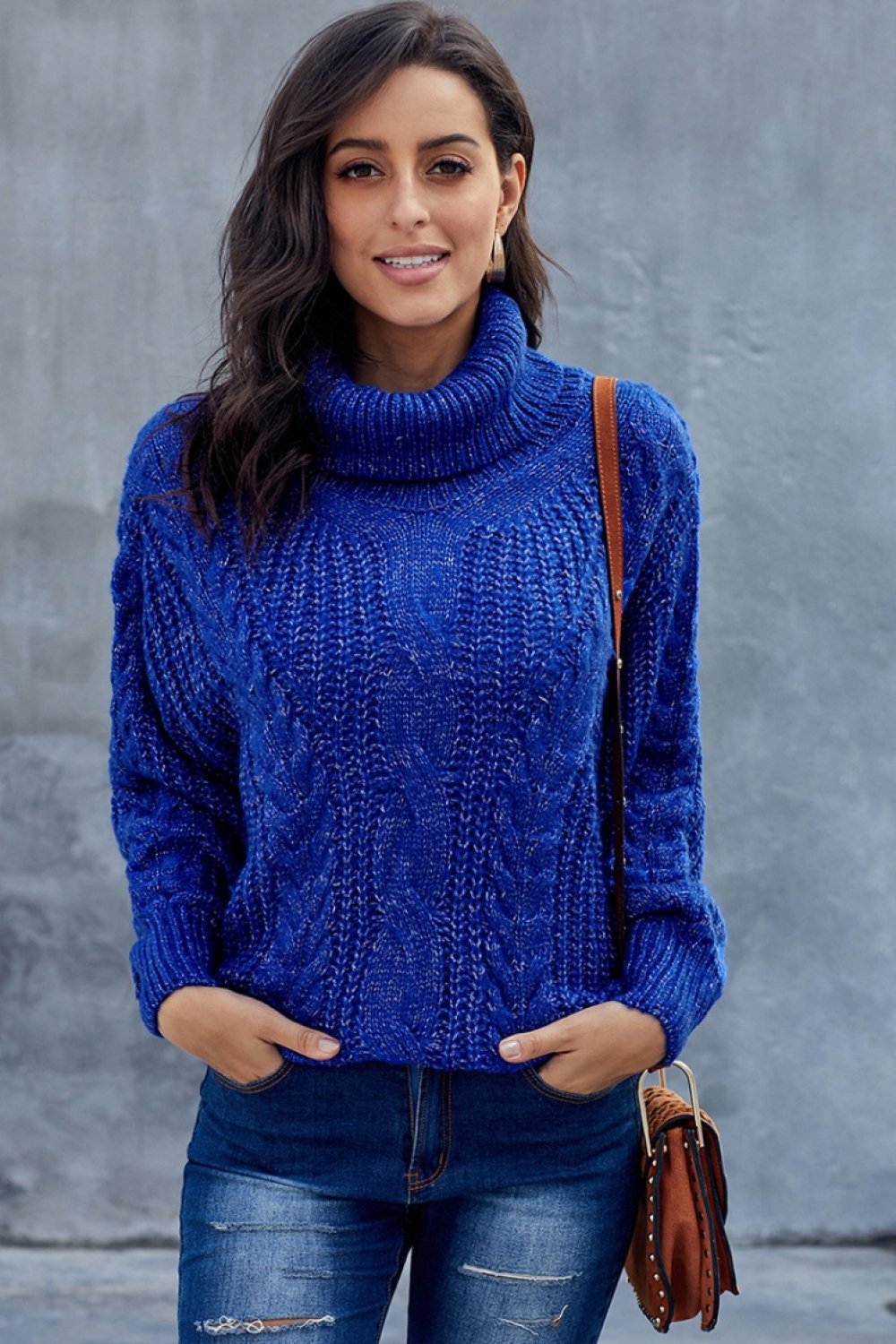 Femme portant un pull bleu à col roulé en tricot torsadé, posant devant un mur gris. Mode automne, style décontracté, tendance féminine.