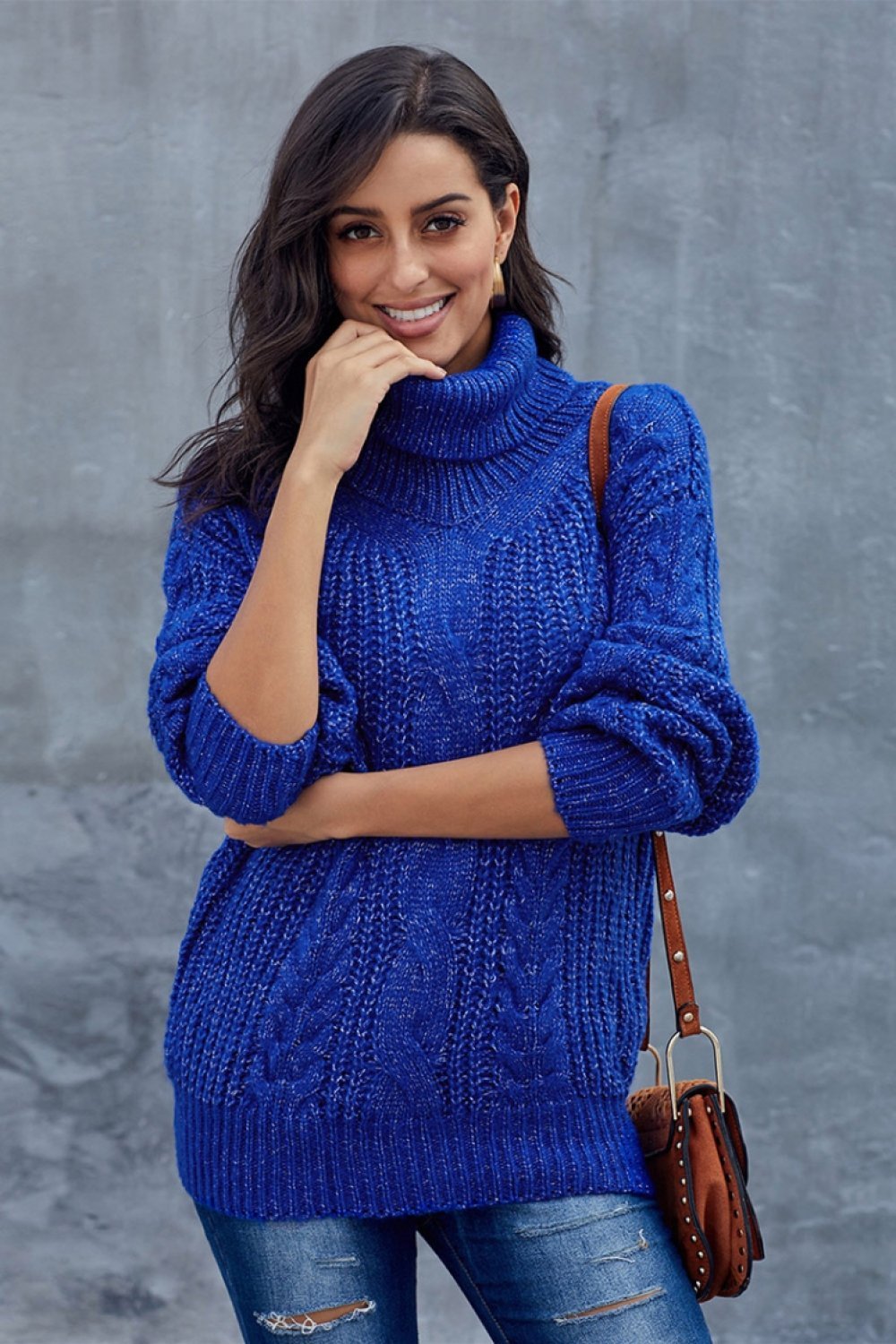 Femme souriante portant un pull bleu en tricot torsadé, manches retroussées, posant devant un mur gris, sac en cuir marron à l'épaule.