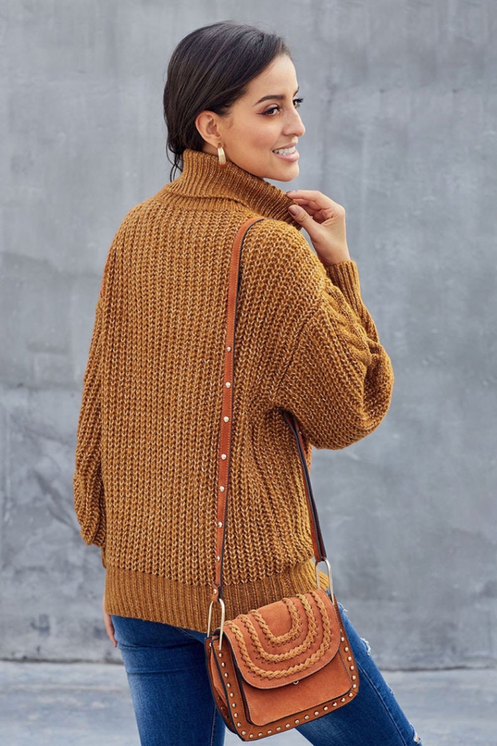 Femme portant un pull en maille marron, sac à bandoulière en cuir, posant de dos. Mode automne, style décontracté, tendance féminine.