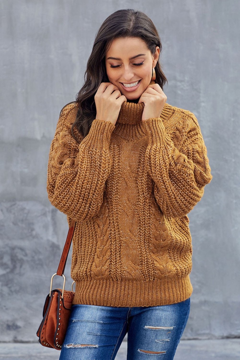 Femme souriante portant un pull en maille torsadée marron, posant devant un mur gris. Mode automne, style décontracté, tendance hiver.