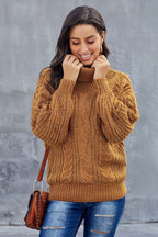 Femme souriante portant un pull en maille torsadée marron, posant devant un mur gris. Mode automne, style décontracté, tendance hiver.