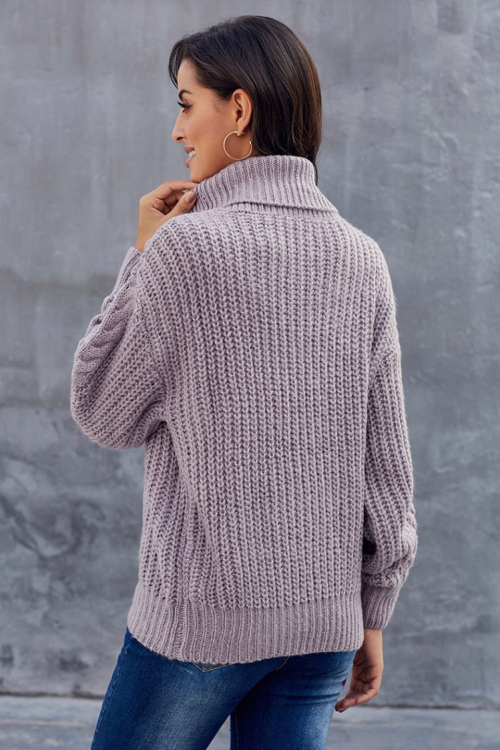 Femme portant un pull en tricot gris à col roulé, manches longues, de dos, devant un mur gris. Mode automne-hiver, style décontracté.