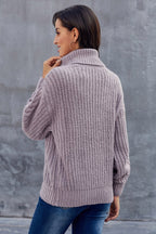 Femme portant un pull en tricot gris à col roulé, manches longues, de dos, devant un mur gris. Mode automne-hiver, style décontracté.