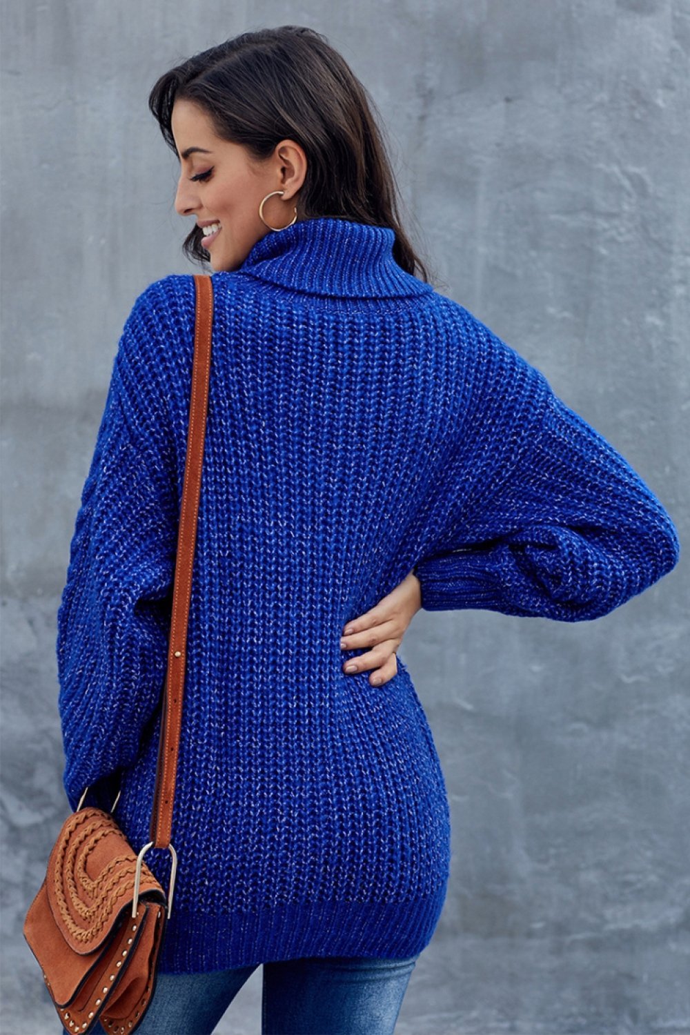 Femme portant un pull bleu en tricot épais, col roulé, avec un sac en cuir marron. Mode automne-hiver, style décontracté et élégant.