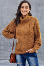Femme souriante portant un pull en maille torsadée marron, sac à main en cuir marron, posant devant un mur gris. Mode automne-hiver décontractée.