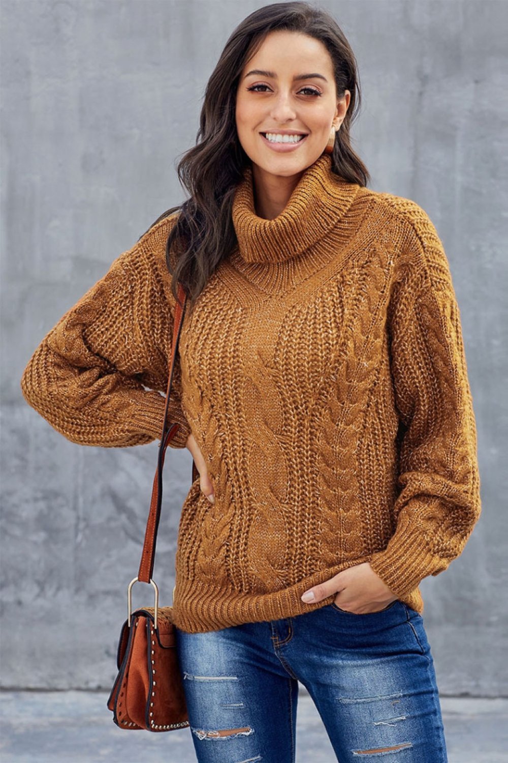 Femme souriante portant un pull en maille torsadée marron, sac à main en cuir marron, posant devant un mur gris. Mode automne-hiver décontractée.