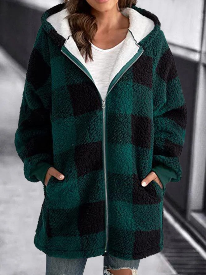 Femme portant un manteau à carreaux vert et noir, style décontracté, avec capuche et poches, sur fond urbain flou. Mode automne-hiver tendance.