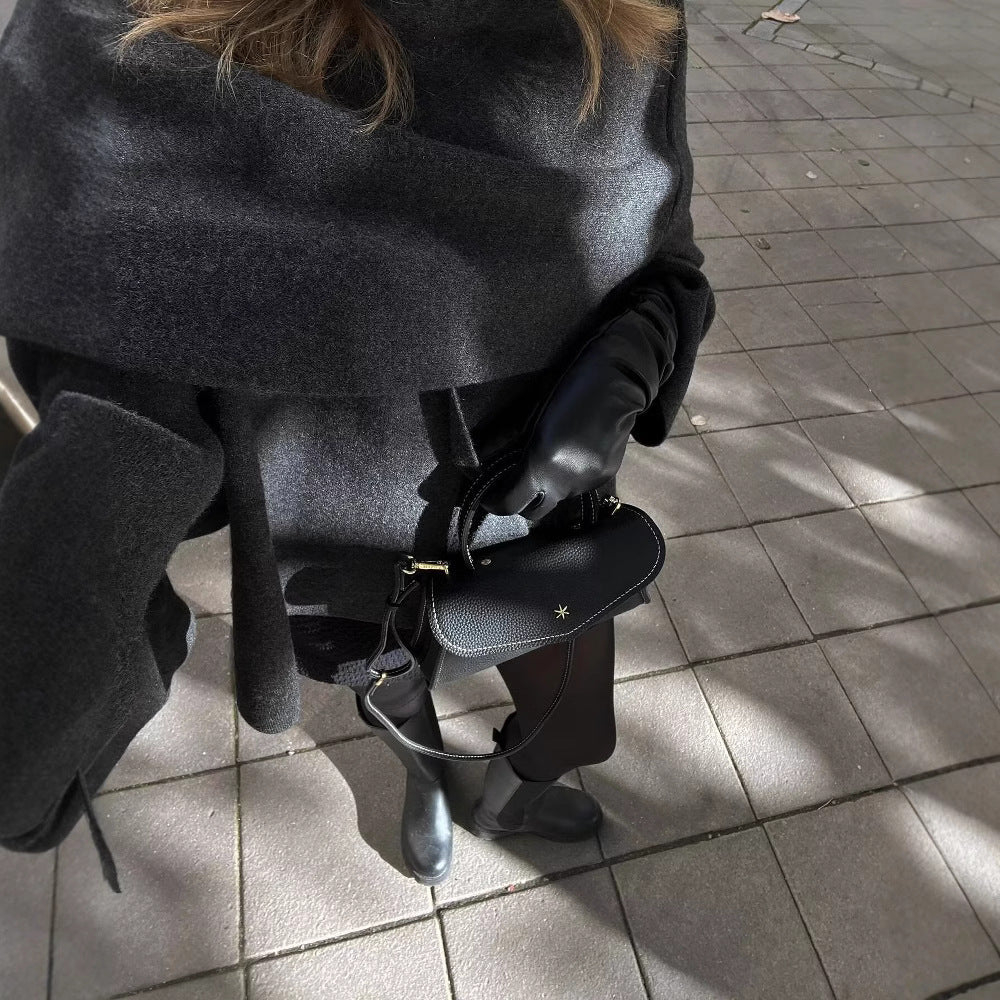 Femme en manteau gris et gants noirs, tenant un sac à main noir, marchant sur un trottoir pavé. Mode automne, élégance urbaine.