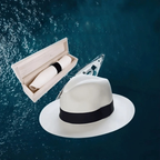 Chapeau Panama blanc avec ruban noir flottant sur fond océan, boîte en bois ouverte à côté. Accessoire élégant, mode estivale, style classique.