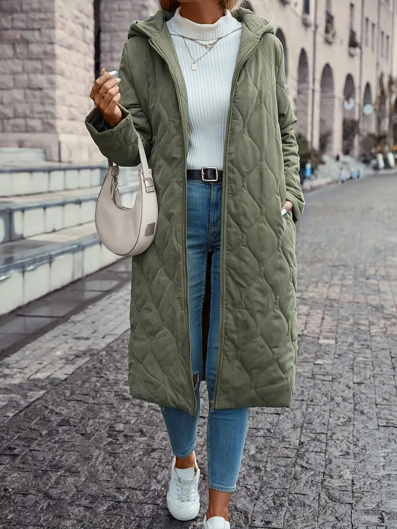 Femme portant un manteau long matelassé vert, jean bleu, pull blanc, sac à main beige, baskets blanches, marchant en ville. Mode automne-hiver tendance.