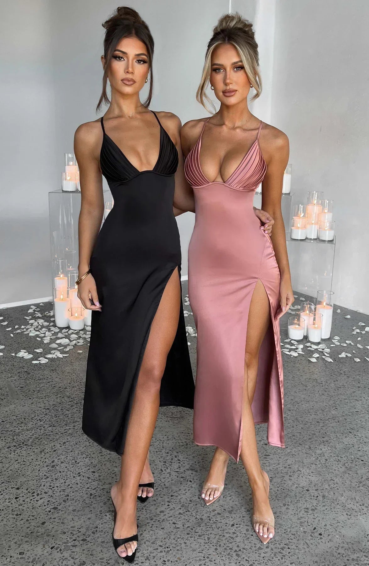 Deux femmes portant des robes longues élégantes, l'une noire et l'autre rose, avec des fentes hautes, posant dans un intérieur moderne avec bougies.