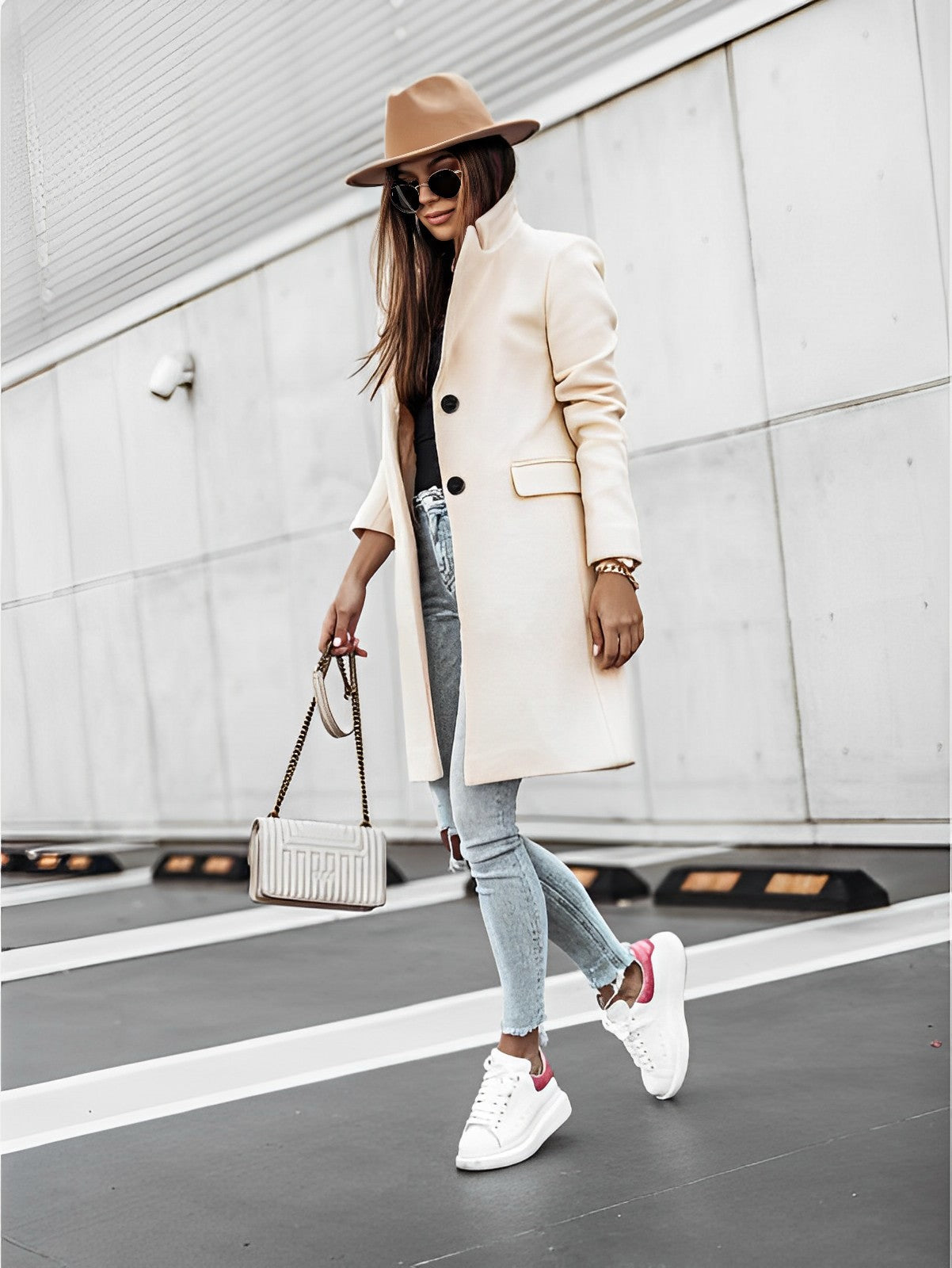 Femme élégante en manteau beige, chapeau marron, jeans déchiré et baskets blanches, portant un sac à main, marchant en extérieur urbain.