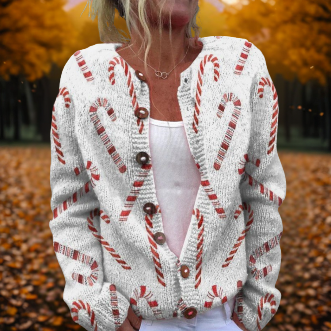 Femme portant un cardigan blanc à motifs de cannes de bonbon rouges, debout dans un parc automnal avec feuilles orange et arbres en arrière-plan.