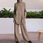 Femme portant un ensemble pull et pantalon beige en laine, posant devant un mur blanc avec des plantes vertes. Mode automne-hiver, tenue confortable.