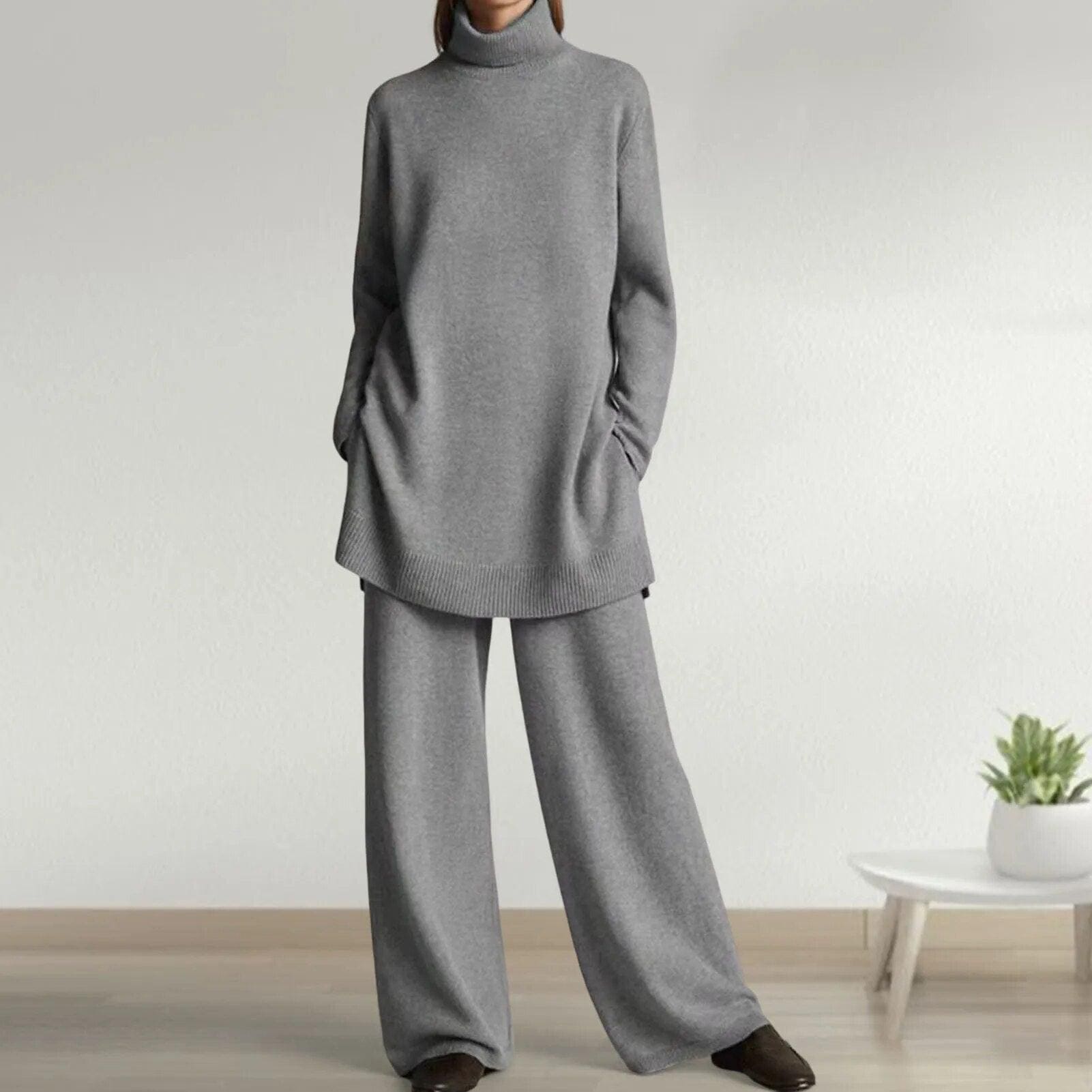 Femme portant un ensemble pull et pantalon gris ample, col roulé, manches longues, dans un intérieur moderne avec plante verte sur table basse.
