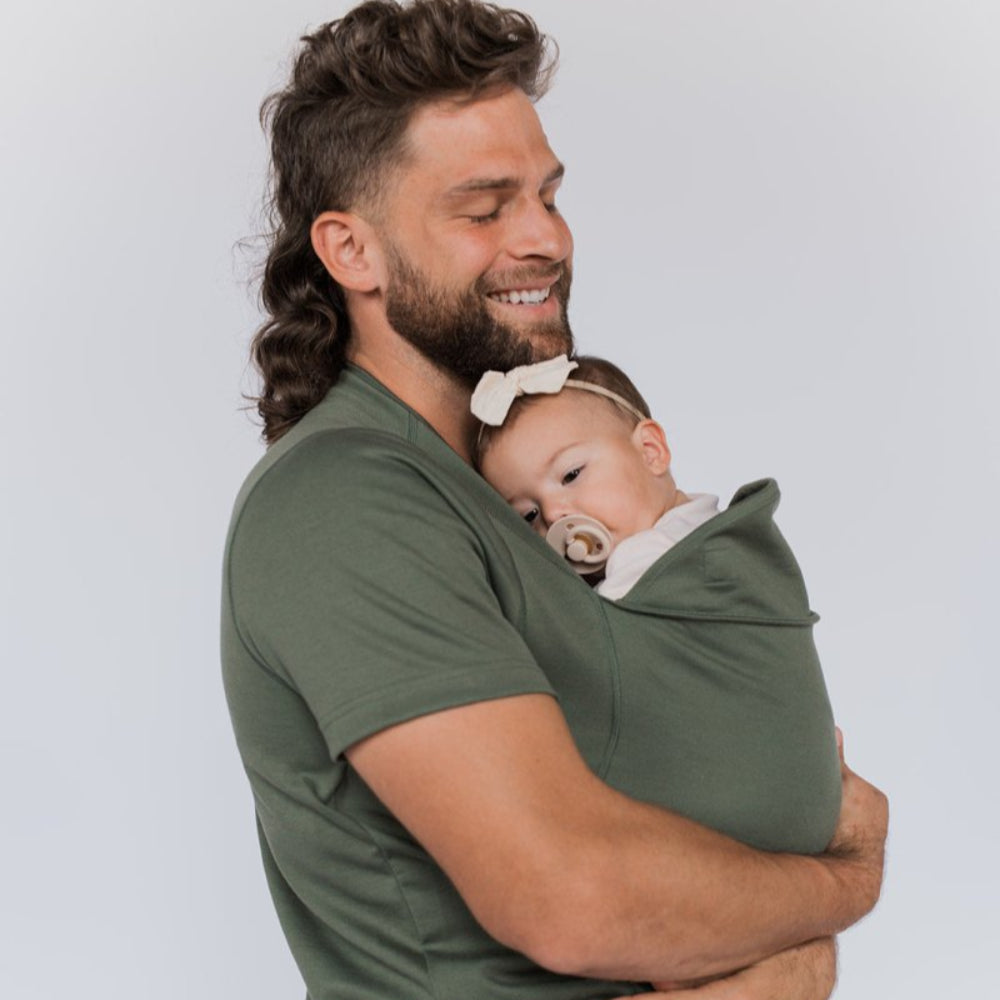 Homme souriant portant un bébé endormi dans un porte-bébé vert, bébé avec tétine et bandeau blanc. Confort, parentalité, proximité.