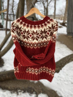 Pull en laine rouge avec motifs nordiques blancs et marron, suspendu à un cintre en bois, fond de neige et arbres d'hiver. Mode hiver chaud.