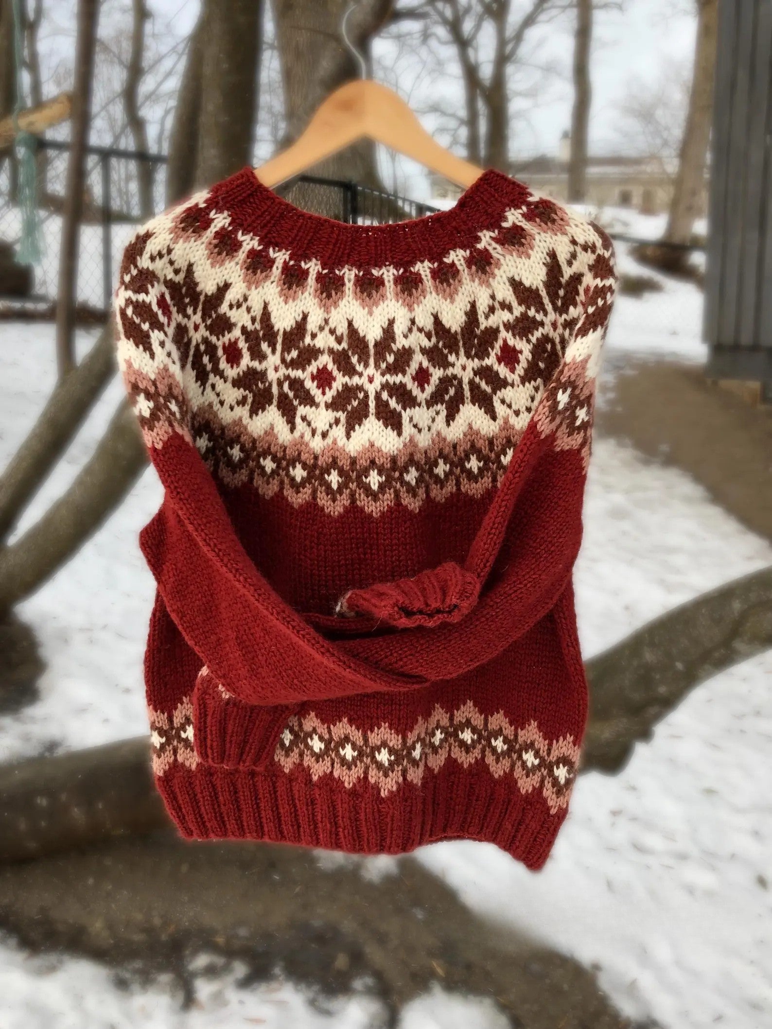 Pull en laine rouge avec motifs nordiques blancs et marron, suspendu à un cintre en bois, fond de neige et arbres d'hiver. Mode hiver chaud.