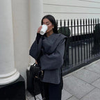 Femme buvant un café à emporter, vêtue d'un manteau gris élégant, debout devant une clôture en fer forgé. Mode automne, style urbain chic.