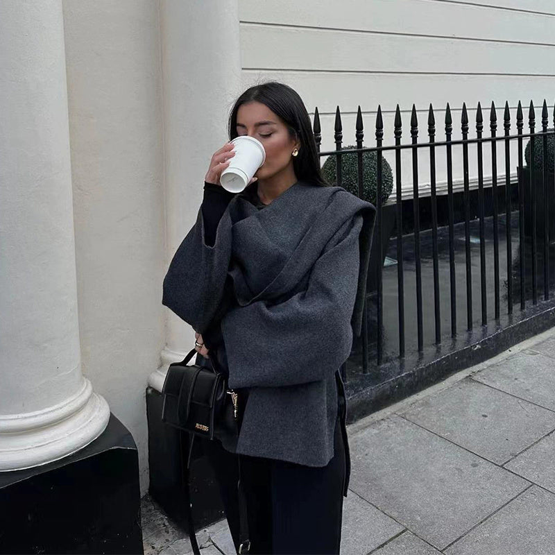 Femme buvant un café à emporter, vêtue d'un manteau gris élégant, debout devant une clôture en fer forgé. Mode automne, style urbain chic.