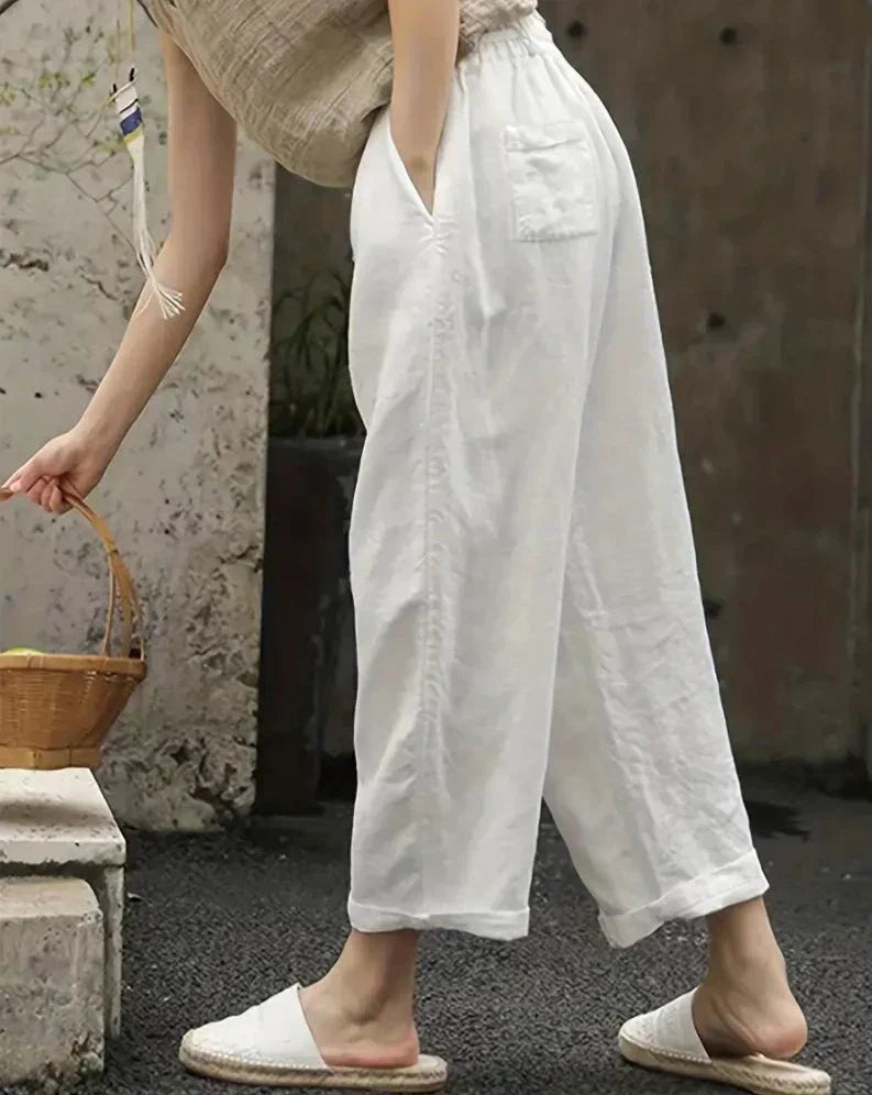 Femme portant un pantalon blanc ample en lin, sandales en toile, tenant un panier en osier. Mode estivale décontractée, style bohème chic.