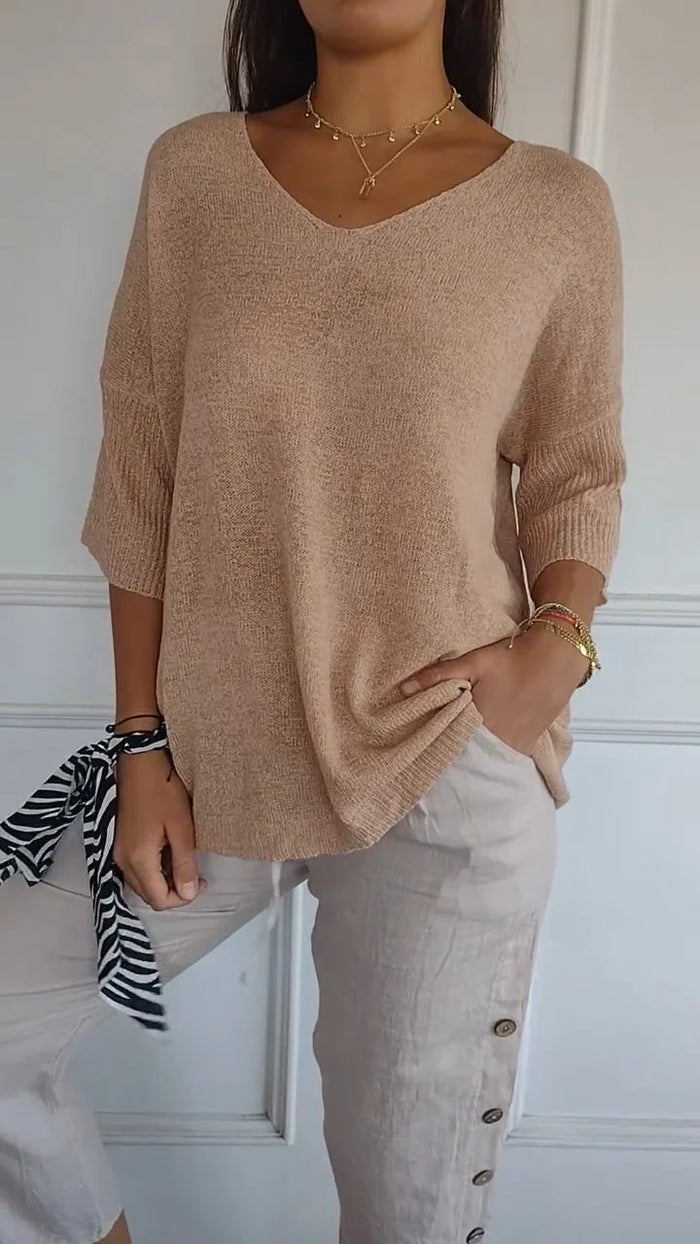 Femme portant un pull beige à col en V, manches trois-quarts, avec un pantalon beige à boutons latéraux. Accessoires: bracelet doré, foulard zébré.