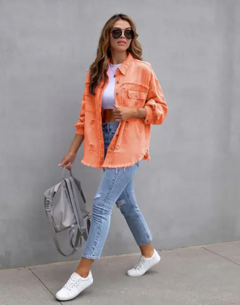 Femme portant une veste orange, jean déchiré, t-shirt blanc et baskets blanches, marchant avec un sac à dos gris. Mode décontractée, tendance urbaine.