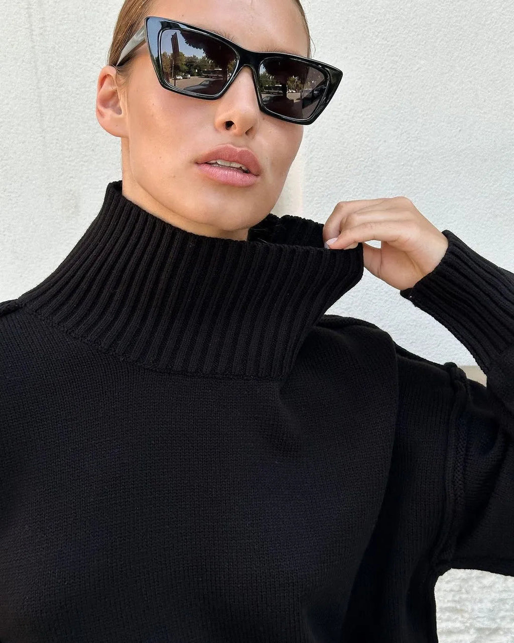Femme portant un pull noir à col roulé, lunettes de soleil tendance, posant devant un mur blanc. Mode automne, style chic et moderne.