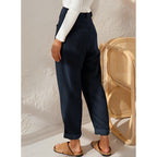 Femme portant un pantalon bleu marine ample, debout près d'une chaise en rotin, sur fond de mur texturé blanc. Mode décontractée élégante.