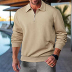 Homme portant un pull beige à col zippé, motif géométrique, près d'une piscine. Mode masculine élégante, tenue décontractée chic.