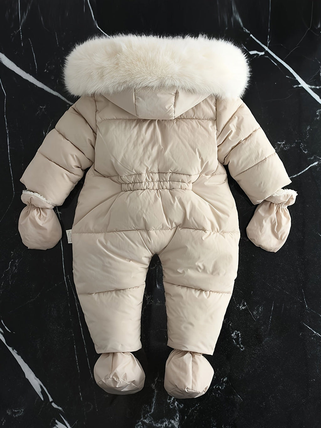 Combinaison bébé en duvet beige avec capuche bordée de fourrure blanche, posée sur fond marbré noir. Vêtement d'hiver chaud et élégant.