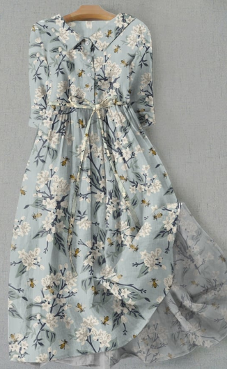 Robe longue fleurie bleue avec motifs floraux blancs et jaunes, manches longues, ceinture à nouer, sur cintre en bois, fond gris.