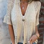 Femme portant un cardigan en maille beige avec un bouton, collier en chaîne argentée et bracelet doré, devant un mur en briques.