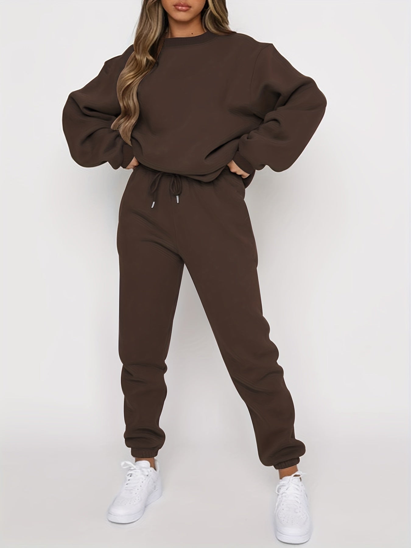 Femme portant un ensemble de survêtement marron, composé d'un sweat ample et d'un pantalon de jogging, avec des baskets blanches. Mode décontractée.