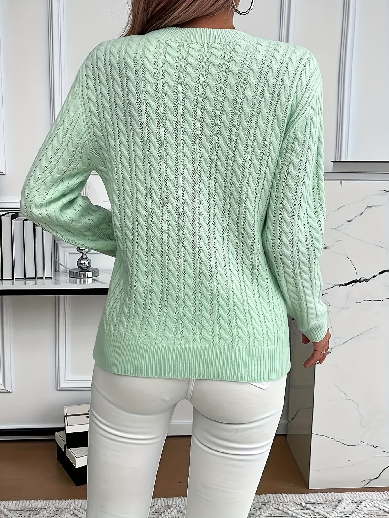 Femme portant un pull torsadé vert menthe, dos tourné, devant une étagère moderne. Mode automne, style décontracté, intérieur élégant.