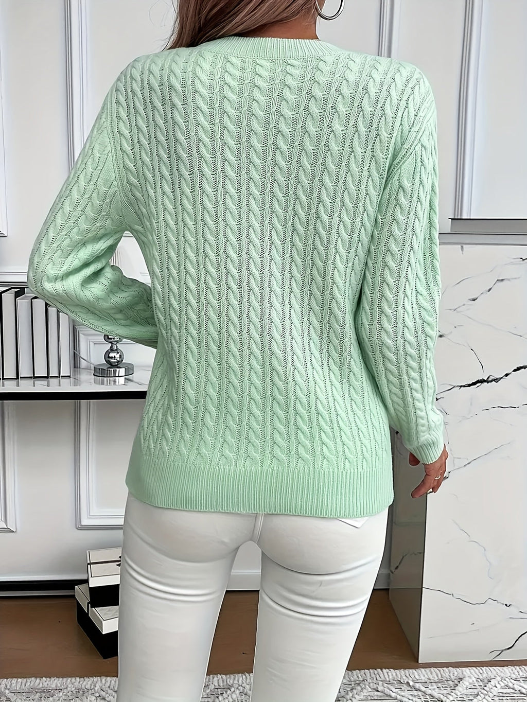 Femme portant un pull torsadé vert menthe, dos tourné, devant une étagère moderne. Mode automne, style décontracté, intérieur élégant.