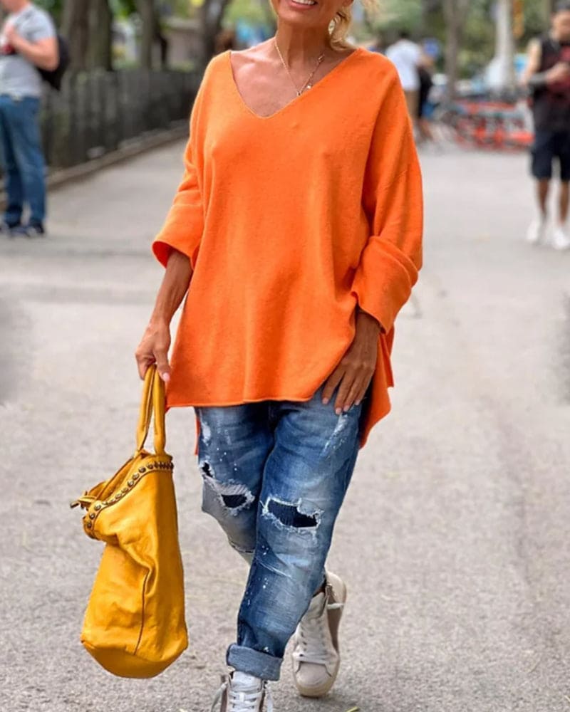 Femme portant un pull orange ample, jean déchiré et baskets blanches, tenant un sac jaune vif, marchant dans une rue urbaine.