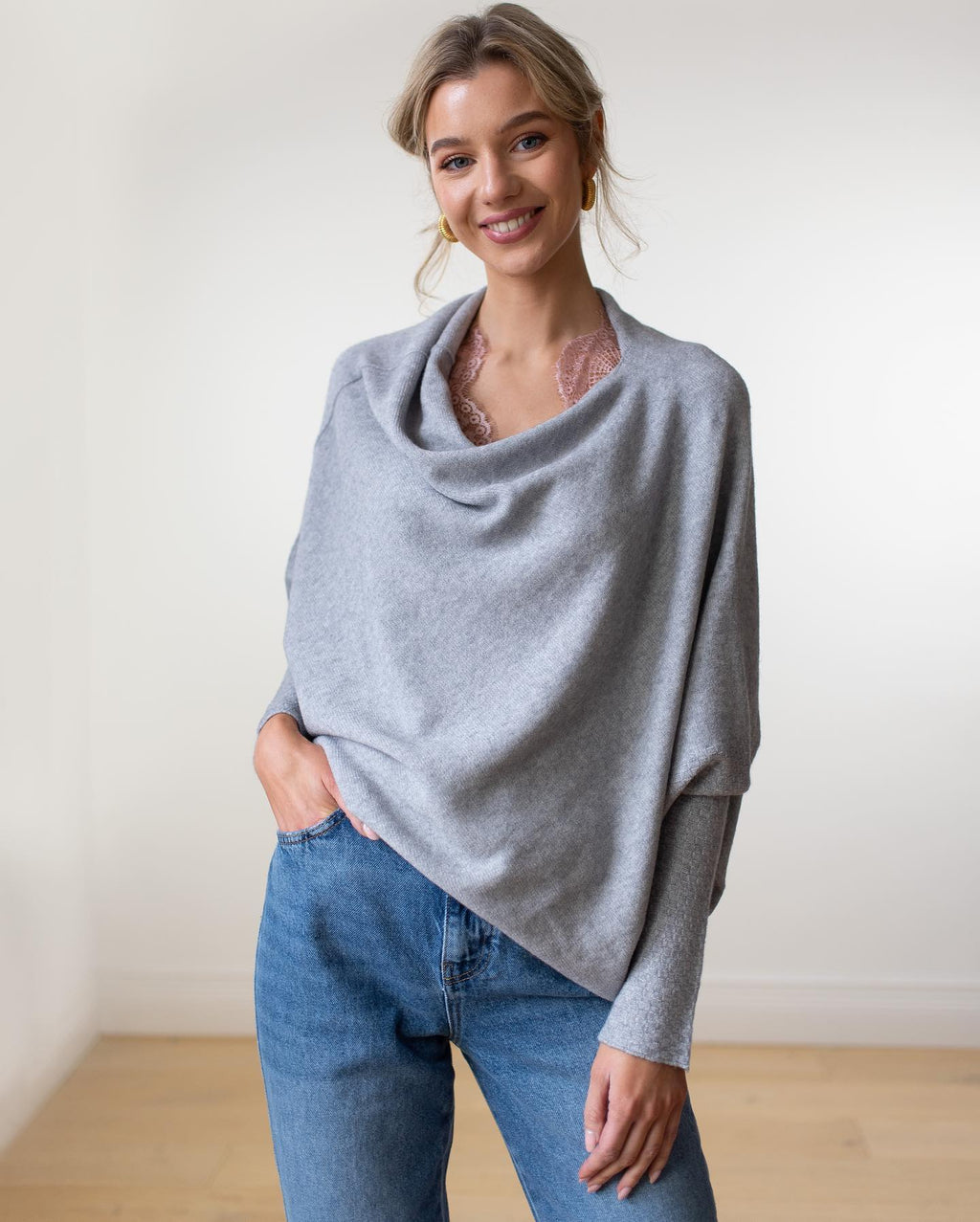 Femme souriante portant un pull gris élégant, col drapé, avec un jean bleu. Mode décontractée, style automne, tenue confortable et chic.