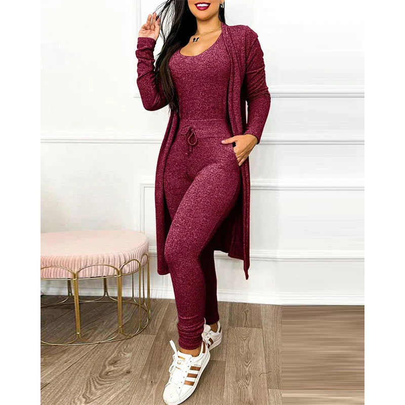 Femme en ensemble bordeaux, combinaison moulante et cardigan long, posant debout. Chaussures blanches, décor intérieur moderne. Mode féminine élégante.