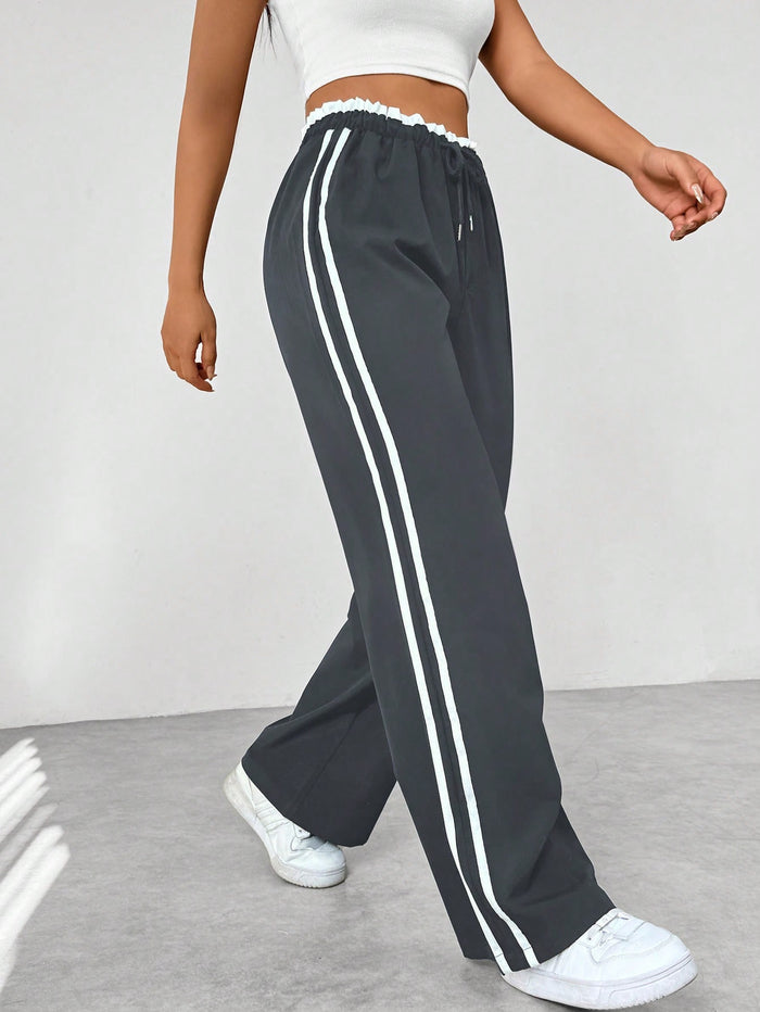 Femme portant un pantalon de jogging noir à rayures blanches, taille élastique, avec un haut blanc, sur fond gris. Mode décontractée et sportive.