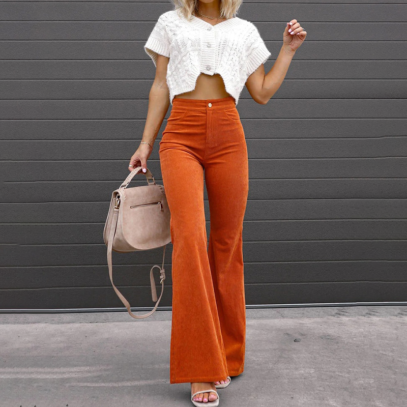 Femme portant un pantalon évasé orange taille haute et un haut court blanc, tenant un sac à main beige devant un mur gris. Mode tendance automne.