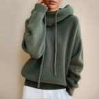 Femme portant un pull à capuche vert olive en laine, manches longues, avec cordons, sur fond beige. Mode automne-hiver, style décontracté.