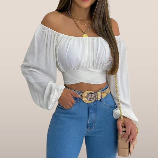 Femme portant un haut blanc à épaules dénudées et manches longues, avec un jean bleu taille haute et une ceinture dorée, mode tendance chic.