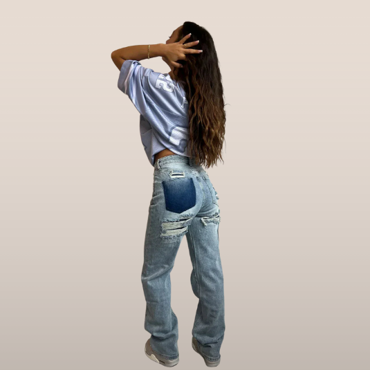 Jeune femme de dos portant un jean déchiré et un t-shirt ample, posant avec style. Mode décontractée, tendance urbaine, cheveux longs ondulés.