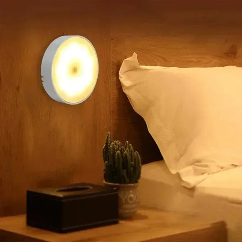 Lampe murale LED ronde émettant une lumière chaude, installée dans une chambre moderne avec un lit et une table de chevet en bois.