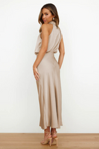 Femme portant une robe longue en satin beige, élégante et fluide, avec des sandales à talons, posant de profil sur un fond blanc minimaliste.