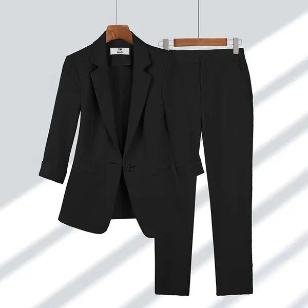 Costume femme noir élégant, veste à manches longues et pantalon assorti, suspendu sur cintres en bois. Mode professionnelle chic.