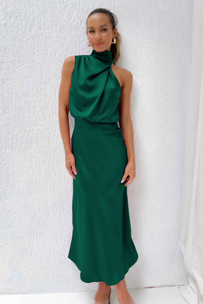 Femme portant une élégante robe longue verte, sans manches, avec un col drapé, posant devant un mur blanc. Mode féminine chic et moderne.