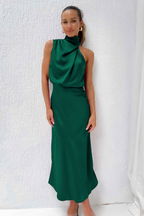 Femme portant une élégante robe longue verte, sans manches, avec un col drapé, posant devant un mur blanc. Mode féminine chic et moderne.